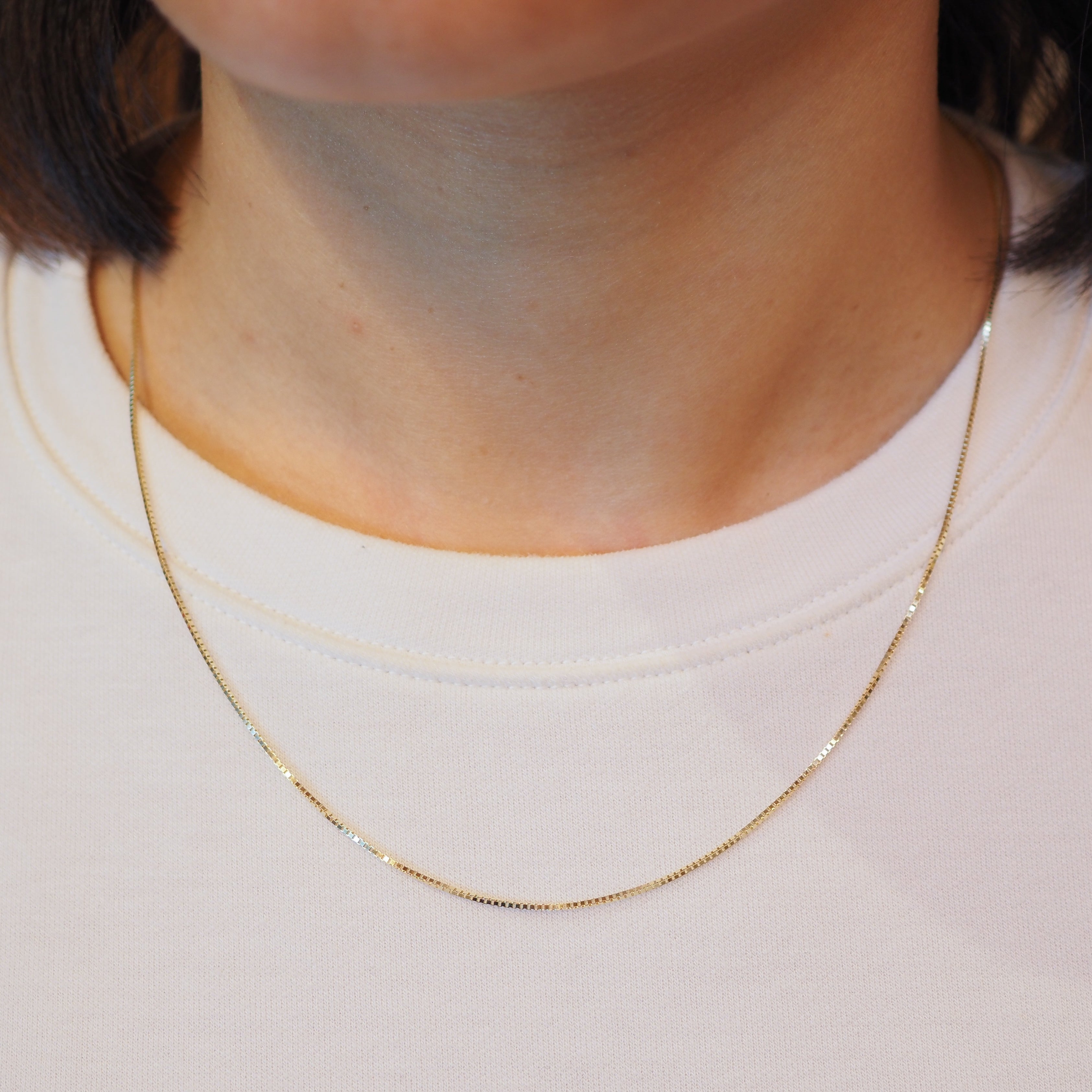 A close-up of a person's neck and upper chest wearing a delicate gold chain necklace over a white crew-neck t-shirt.