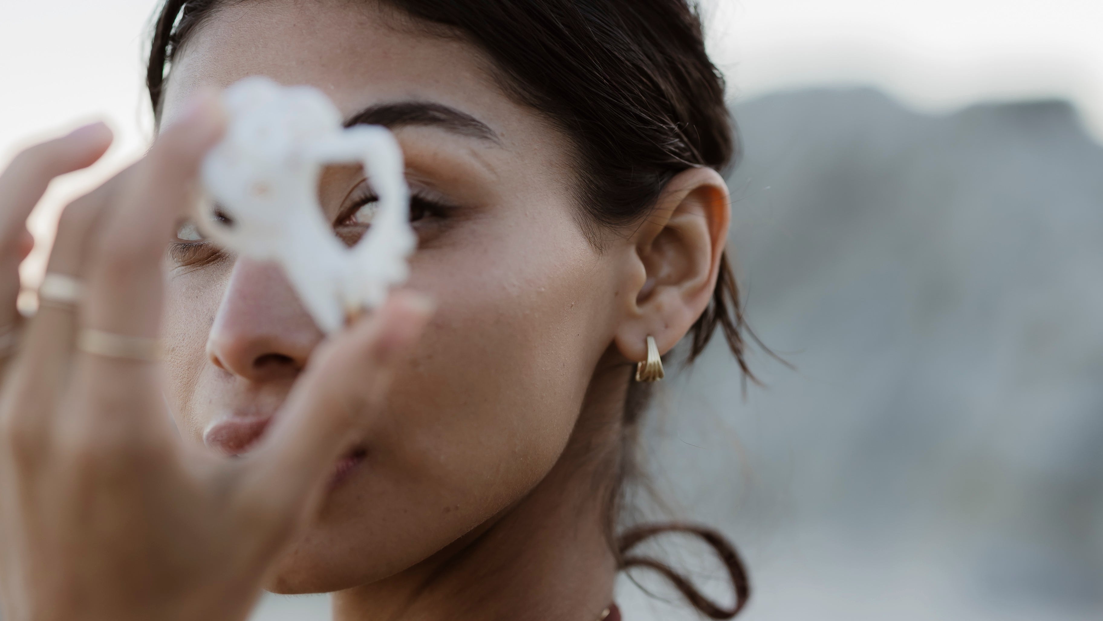 Woman holding a white shell to her face, wearing gold shell huggie hoop earrings with a blurred natural background
