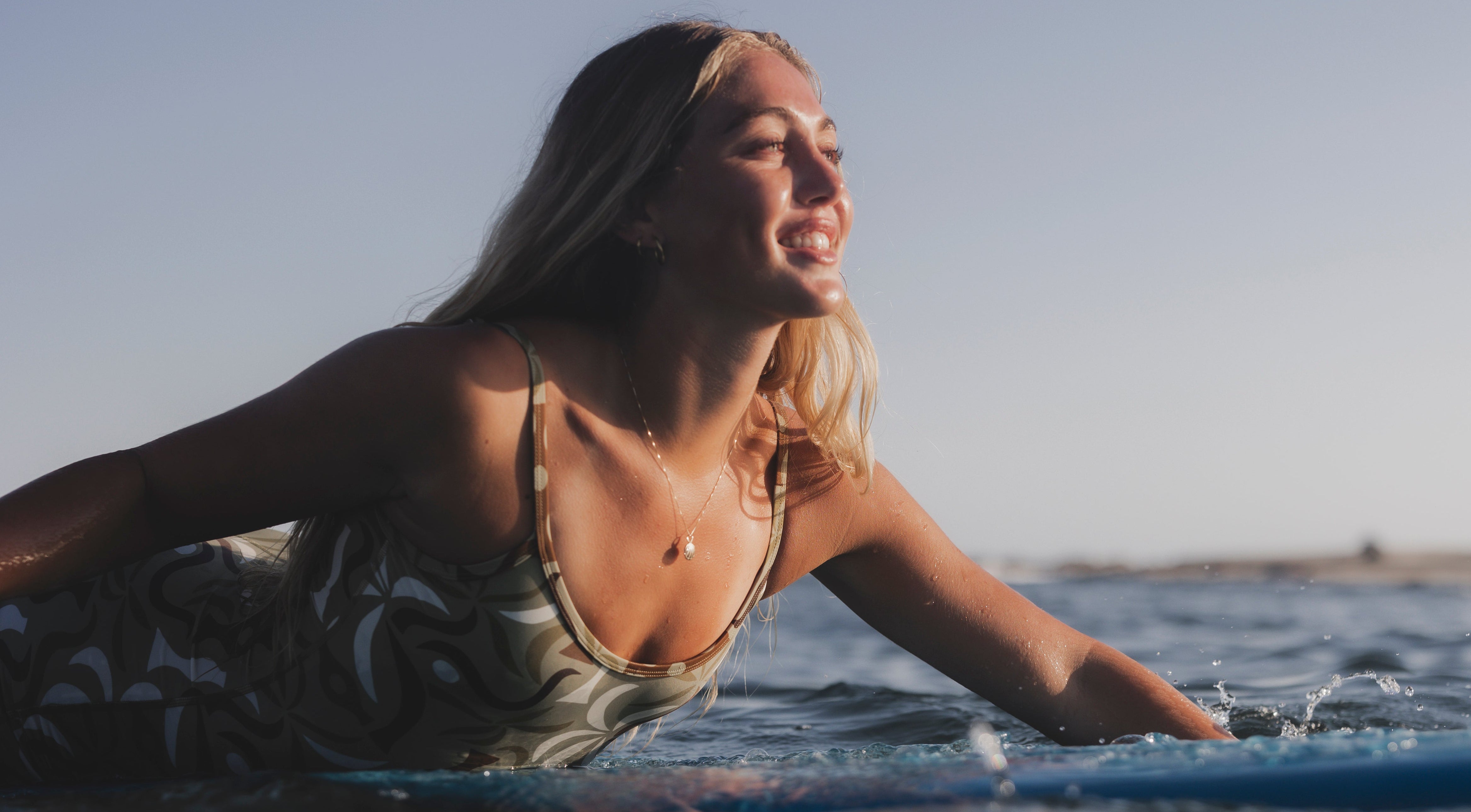 Woman lying on a surfboard wearing a gold shell necklace in the ocean with a clear sky.