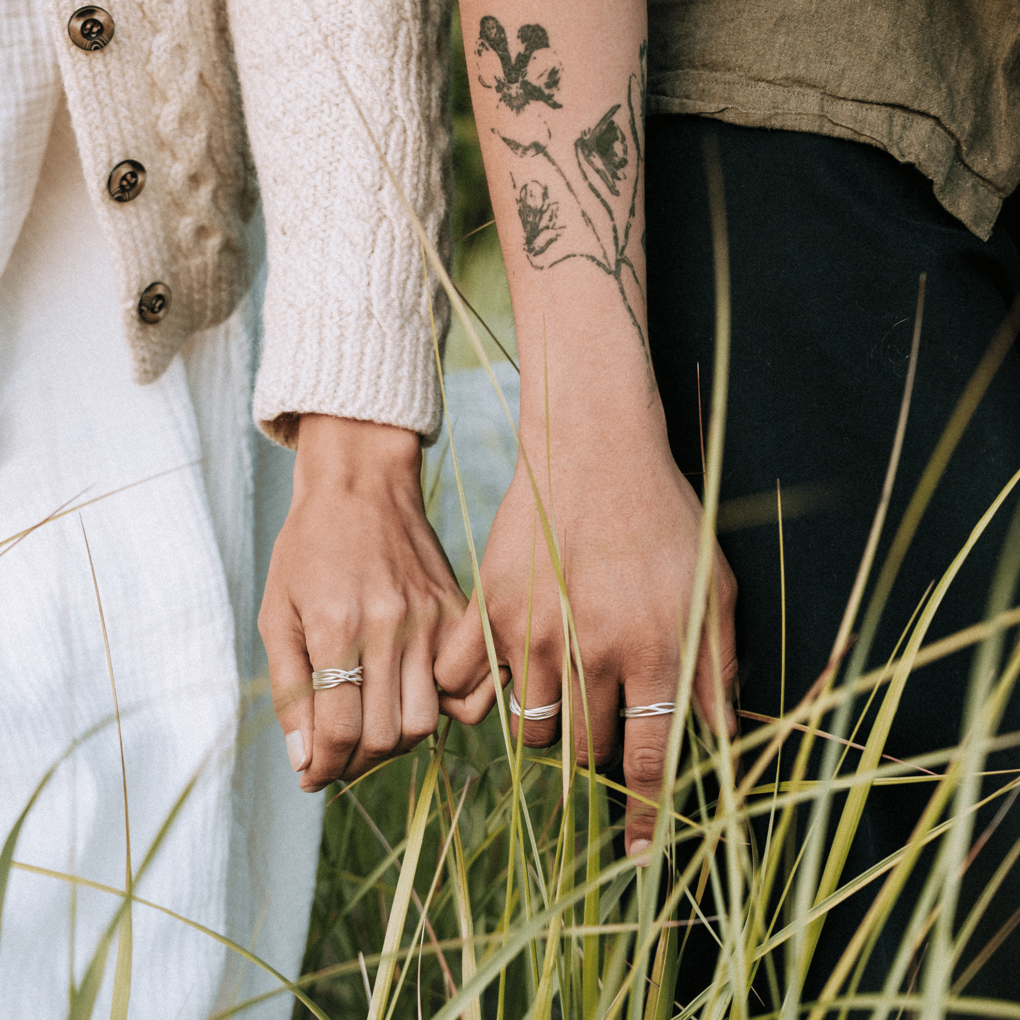 Two people holding hands with tattoos visible, standing in tall grass.