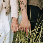 Two people holding hands with tattoos visible, standing in tall grass.